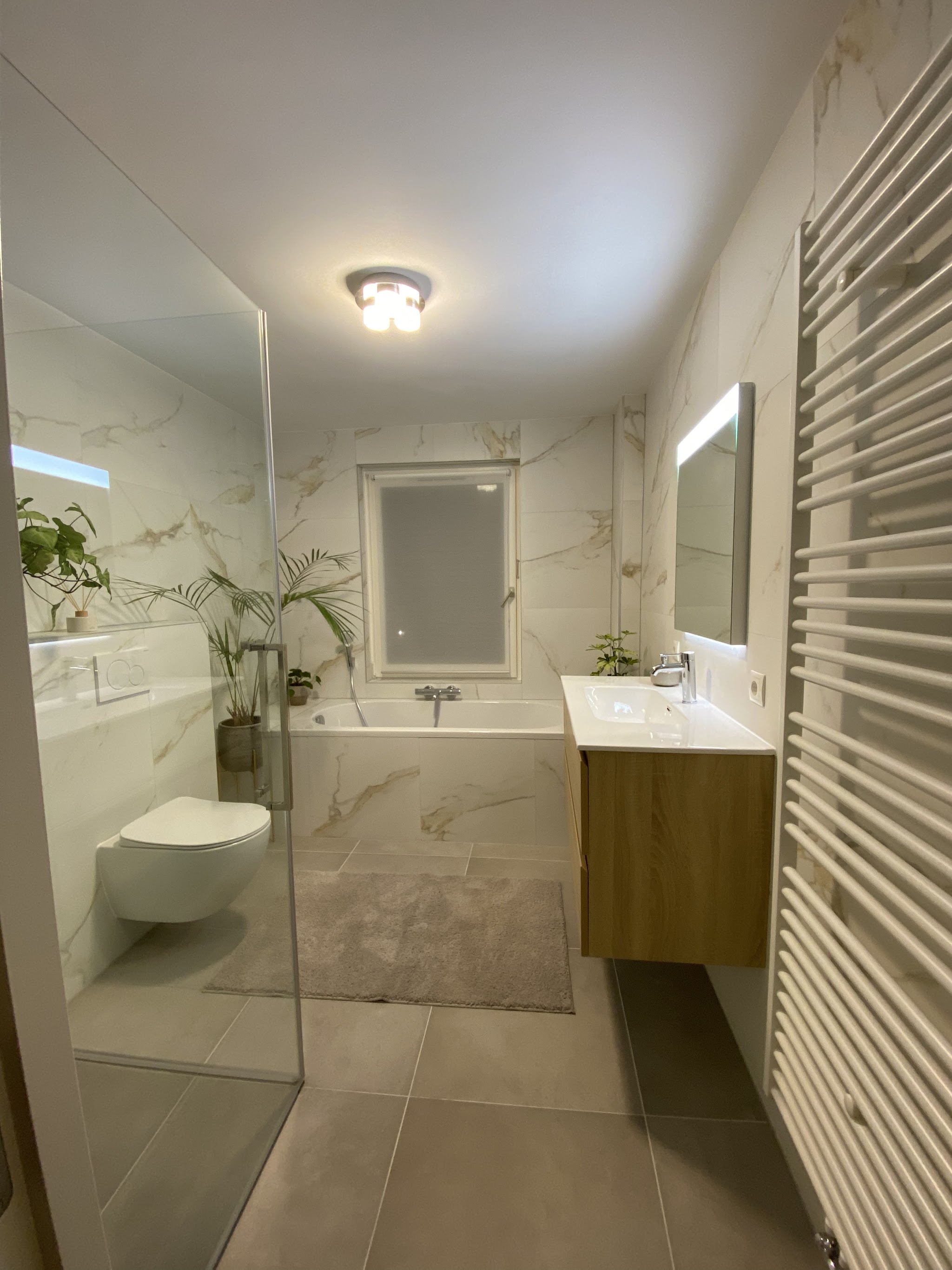 Lichte badkamer met marmerlook tegels, glazen douchewand, ligbad en eikenhouten badmeubel