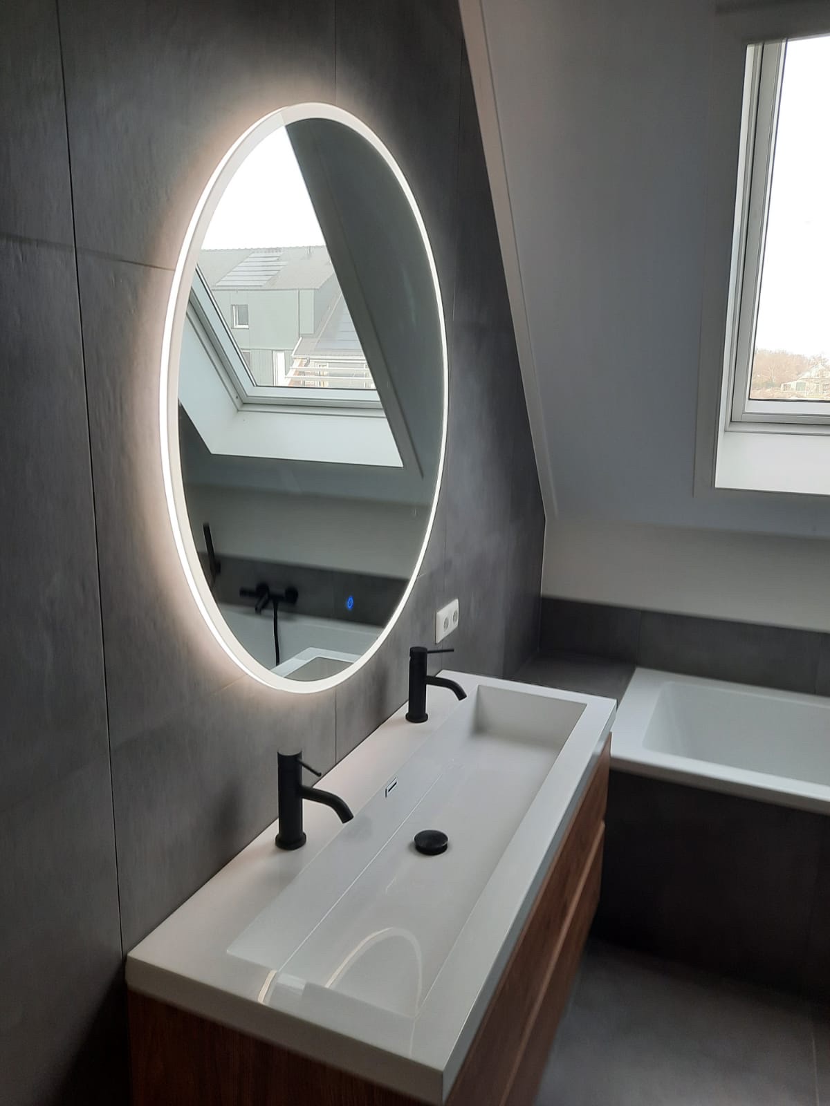 Zolderbadkamer met ronde verlichte spiegel en ligbad onder het dakraam
