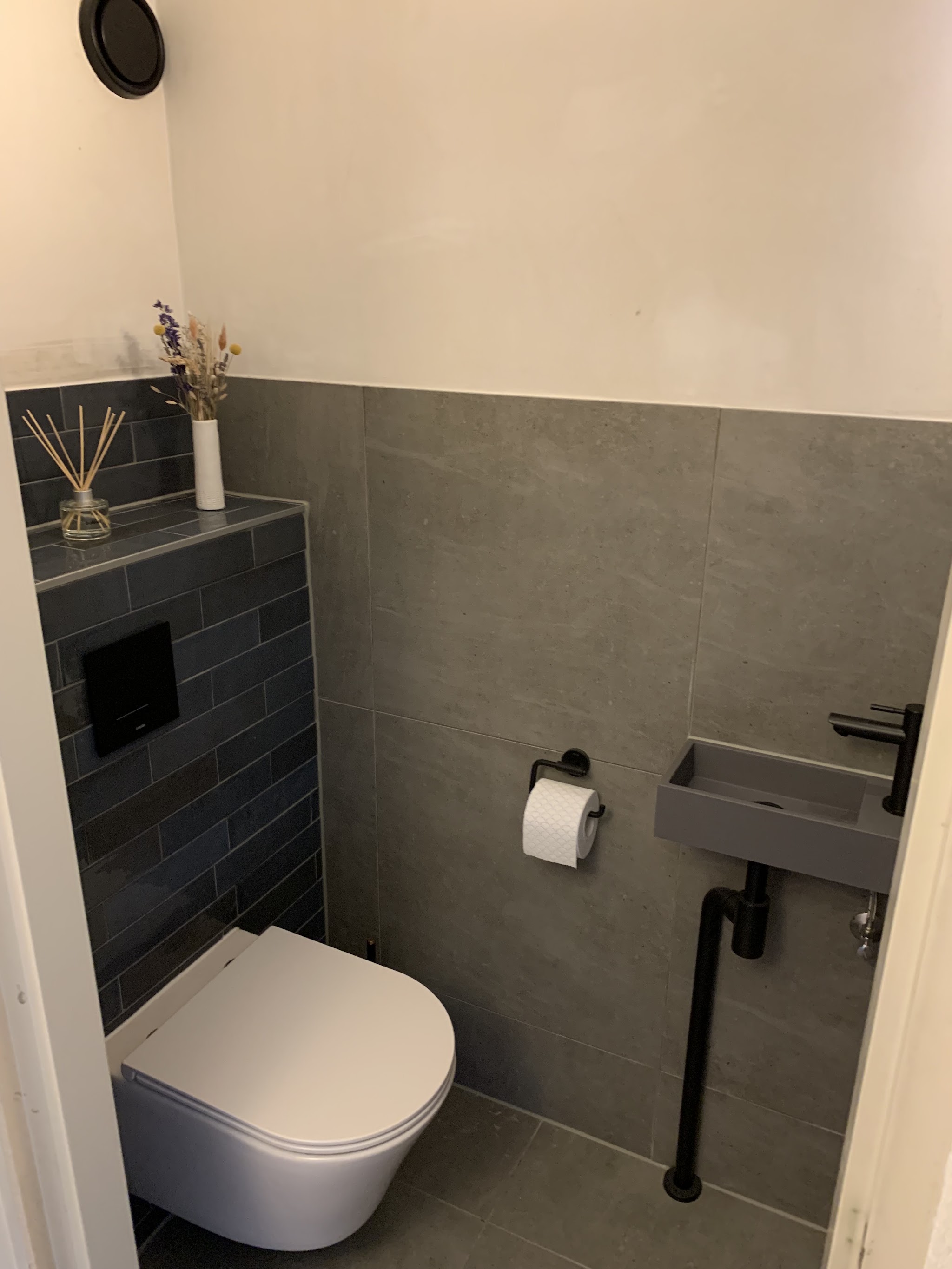 Toiletruimte met donkere tegelwand, hangtoilet en klein grijs fonteintje