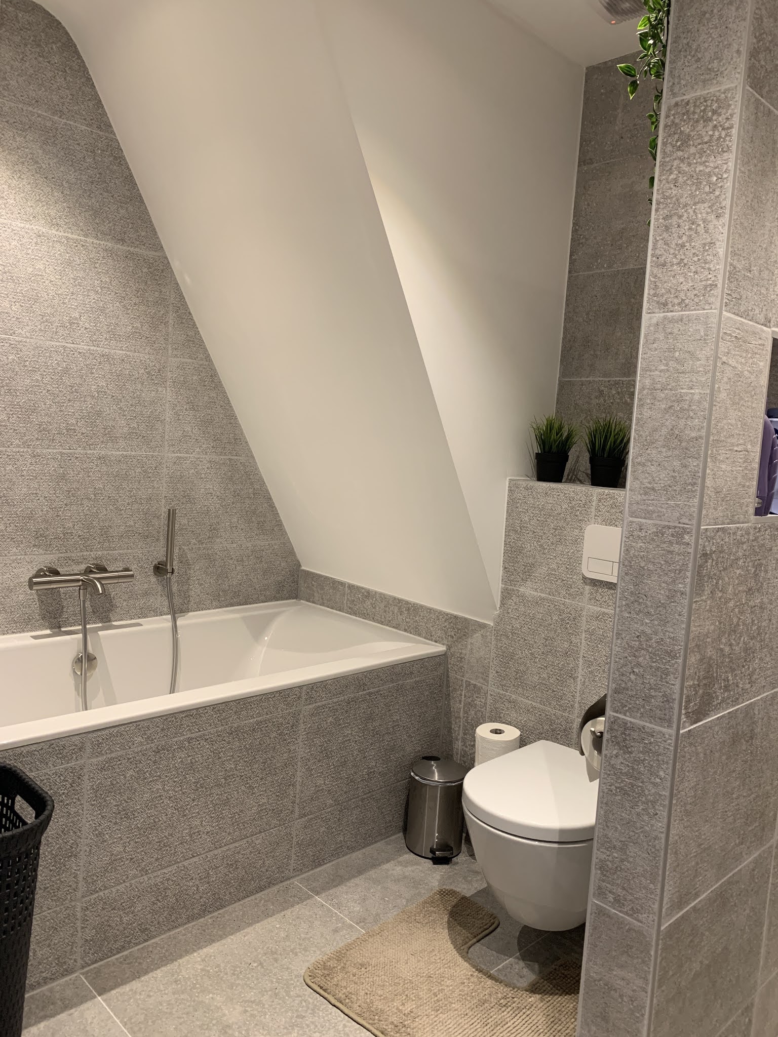 Zolderbadkamer met ligbad en toilet in grijze natuursteenlook tegels